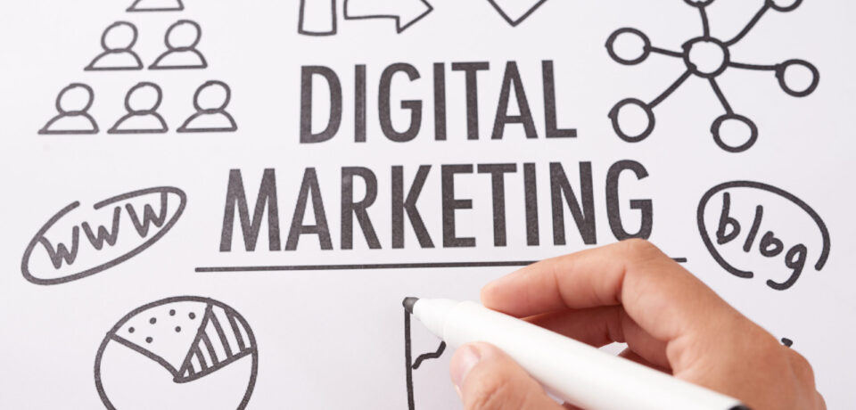 From above shot of crop hand using marker pen to draw icons near digital marketing writing on paper sheet